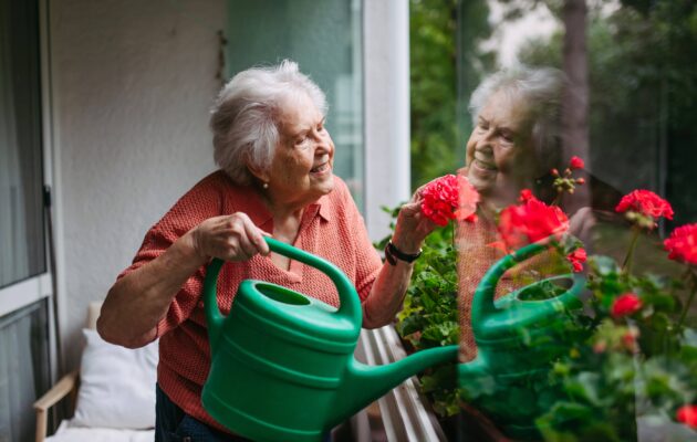 Starejša ženska z demenco zaliva rože na balkonu in ohranja samostojnost, dostojanstvo ter občutek vrednosti pri vsakdanjih opravilih.