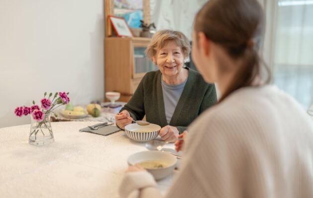 Oseba z demenco sedi za mizo in je juho ob podpori svojke, primer mirnega hranjenja in občutka varnosti pri demenci.