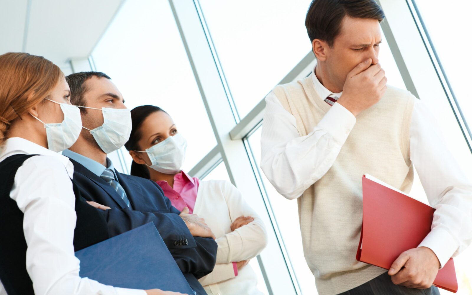 Sodelavci z zaščitnimi maskami stojijo ob moškem, ki kašlja v pisarni, kar ponazarja tveganje okužbe v kolektivu.
