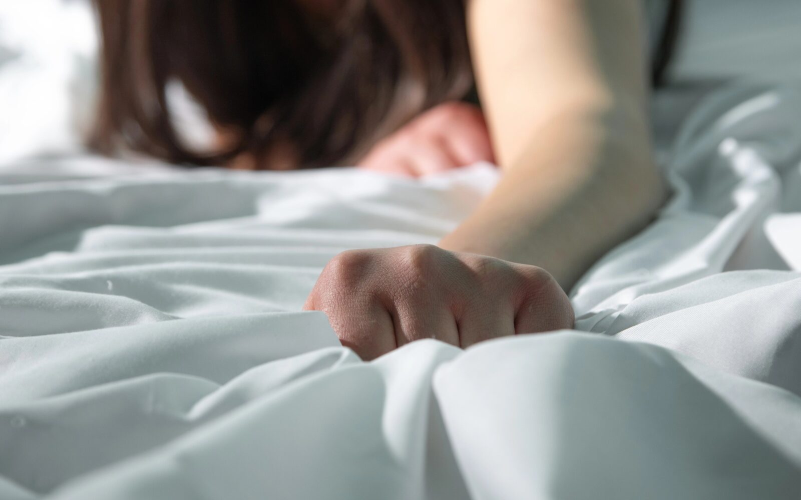 Ženska leži na postelji in z roko "grabi" rjuho, simbolični prikaz doživljanja orgazma