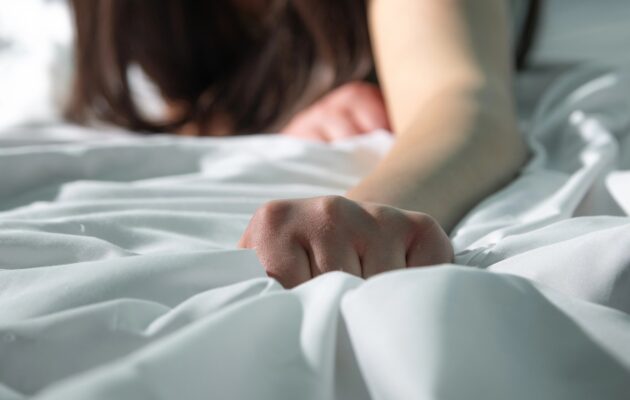 Ženska leži na postelji in z roko "grabi" rjuho, simbolični prikaz doživljanja orgazma