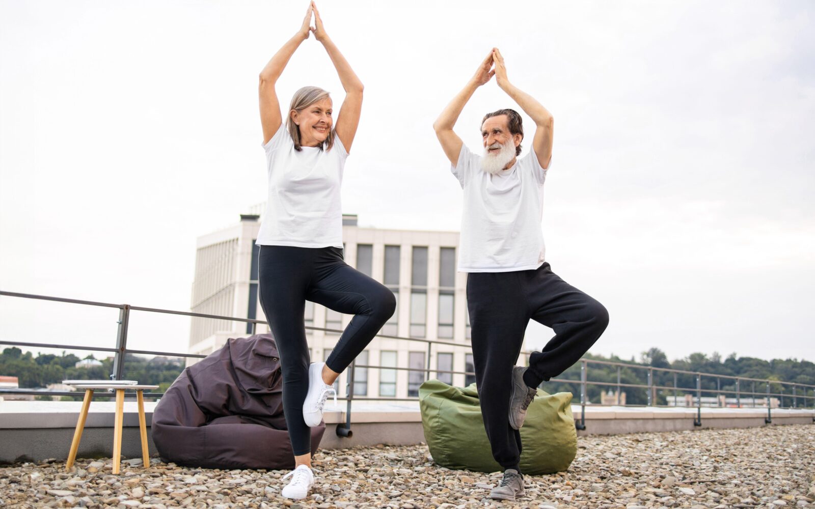 Starejši par na strehi stavbe izvaja jogo - stojita na eni nogi in lovita ravnotežje - znak zdravega staranja