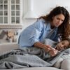 Mama meri telesno temperaturo otroku z vročino, ki leži zavit v odejo na kavču ob okužbi.