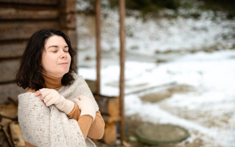 Mlada ženska se zadovoljno smeji in stoji pred hišo na mrazu, zavita v topel šal.