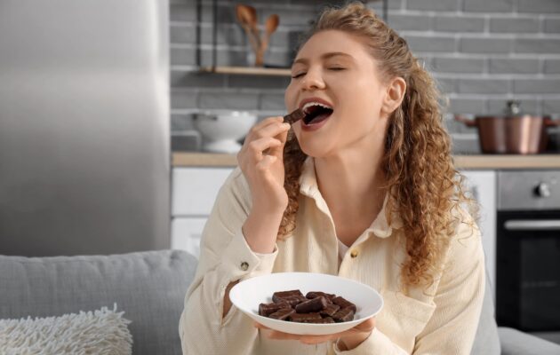 Mlada ženska sedi na kavču in je čokolado - nenaden dvig in padec krvnega sladkorja po neuravnoteževenm obroku